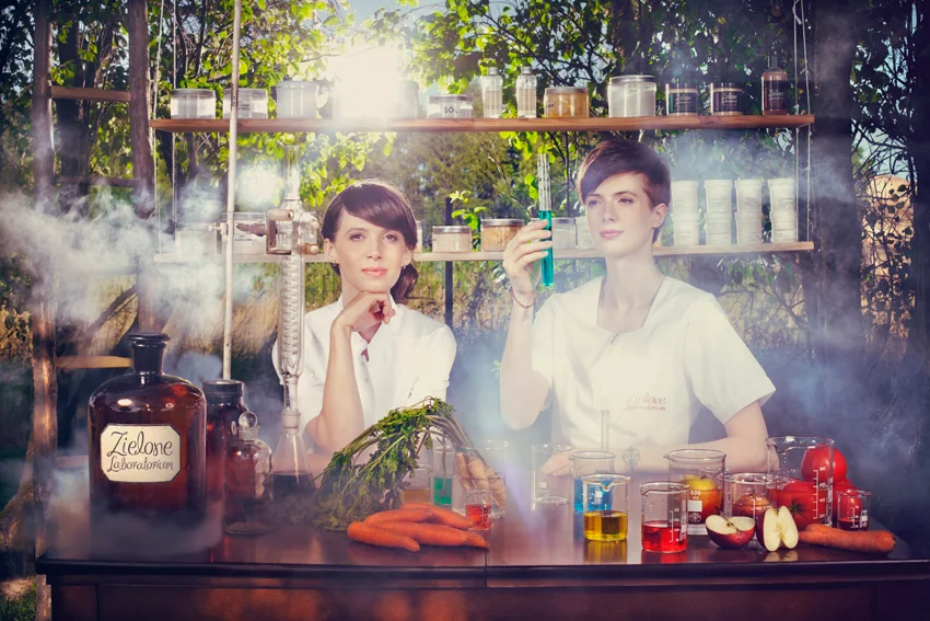Aleksandra i Sylwia Dziegieć w białych fartuchach przy szklanej aparaturze laboratoryjnej, w tle ogród