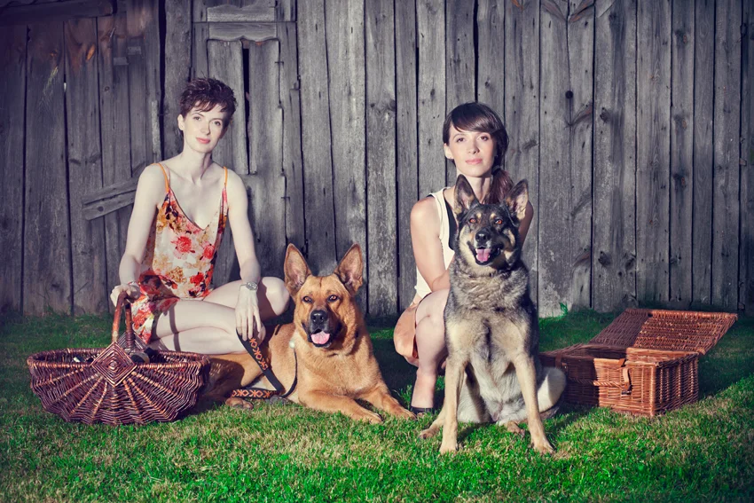 Sylwia i Aleksandra Dziegieć z dwoma psami i wiklinowymi koszami przy drewnianej stodole, 2013